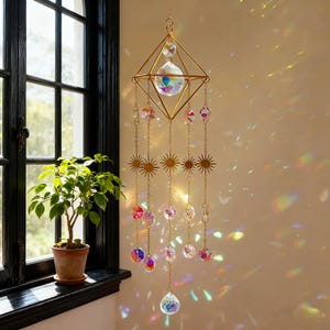 Puede incluir: Un atrapasoles colgante con un marco geométrico dorado y cristales colgantes. El atrapasoles presenta una gran bola de cristal en el centro, con cristales más pequeños y detalles en forma de estrella colgando debajo. Los cristales crean reflejos de luz coloridos.