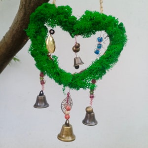 Puede incluir: Una corona de musgo en forma de corazón con campanas y cuentas colgantes. La corona es verde y las campanas son doradas y plateadas.