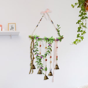 May include: A handmade wall hanging with a wooden branch base, adorned with green leaves, pink and purple flowers, and small bells. The hanging features a variety of beads and a pink ribbon.