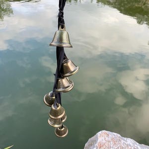 Könnte beinhalten: Eine Ansammlung kleiner, antiker Messingglocken, die an einem schwarzen Band hängen. Die Glocken sind unterschiedlich groß und vor dem Hintergrund eines ruhigen Gewässers mit Himmels- und Wolkenreflexionen angeordnet.