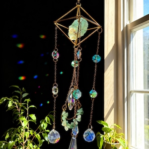 Puede incluir: Un atrapasoles colgante con un marco geométrico dorado que sostiene un cristal verde claro. Cuentas y cadenas de cristal transparentes e iridiscentes cuelgan debajo. El atrapasoles está suspendido frente a una ventana.