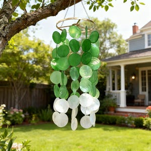 May include: A hanging wind chime with a brass-colored ring and chains. The chime features graduated green and white circular shell-like pieces. The wind chime is suspended from a tree branch in a backyard setting.