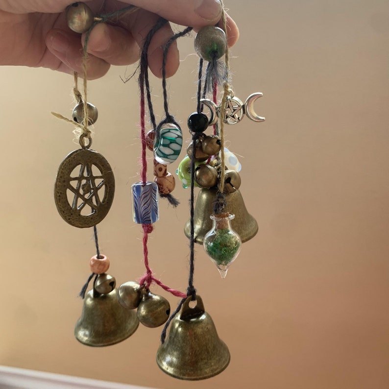 May include: A set of five brass bells with various charms and beads hanging from them. The charms include a pentagram, a crescent moon, and a small glass bottle filled with green glitter. The beads are green, blue, and brown.