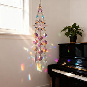 May include: A decorative suncatcher with a gold chain, featuring a starburst design and hanging faceted crystals. The suncatcher has a mix of colorful gems and crystals that create a rainbow effect when light shines through them. It hangs near a window.