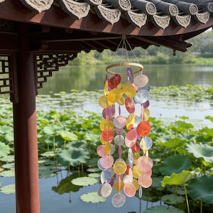 May include: A colorful wind chime with a wooden ring and strands of iridescent, round shell pieces in shades of red, yellow, pink, and blue. The chime hangs from a traditional Asian-style structure with a lake in the background.
