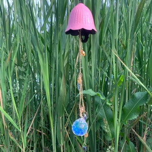 May include: A pink bell-shaped wind chime with a black rim and a gold chain. The chain is adorned with small, clear and amber-colored beads, and a large, iridescent crystal ball. The chime is suspended against a backdrop of tall green reeds.