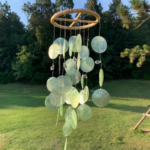 Peut inclure: Un carillon éolien suspendu, composé d'un anneau en bois et de disques verts translucides de différentes formes. Des perles de cristal transparentes sont attachées aux fils. Le carillon est suspendu à l'extérieur, sur fond d'arbres et d'herbe vertes.