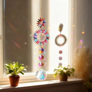May include: A decorative suncatcher with a gold chain, featuring a starburst and a circular design, both adorned with colorful crystals. Below, a series of faceted crystals lead to a large, iridescent crystal ball. The suncatcher hangs in front of a window.