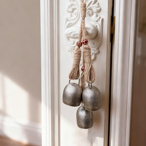 Peut inclure: Trois cloches argentées à surface texturée suspendues à une corde naturelle. La corde est ornée de petites perles rouges et marron. Les cloches sont suspendues à une porte blanche avec des moulures décoratives.