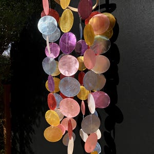 May include: A colorful wind chime with iridescent, round, and oval-shaped shell pieces in shades of pink, purple, yellow, and blue. The chime hangs from a wooden ring, creating a decorative and eye-catching display.