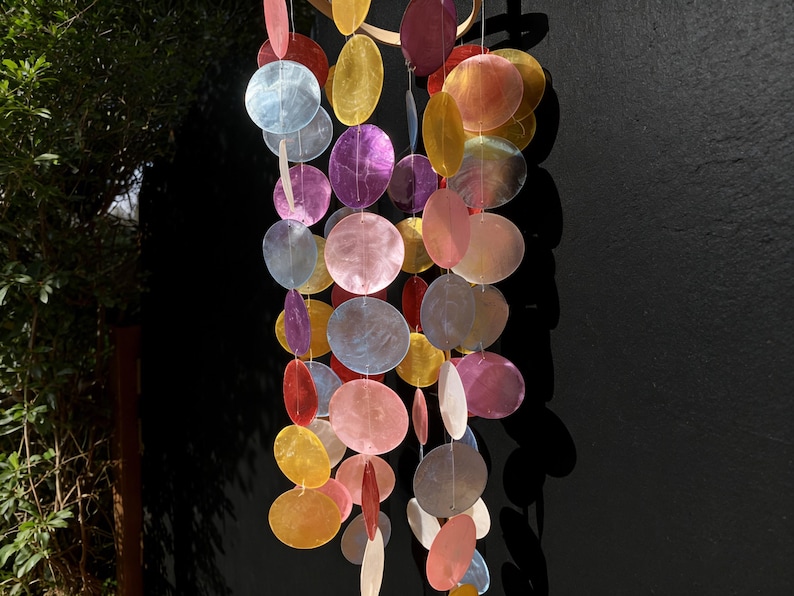 May include: A colorful wind chime with iridescent, round, and oval-shaped shell pieces in shades of pink, purple, yellow, and blue. The chime hangs from a wooden ring, creating a decorative and eye-catching display.