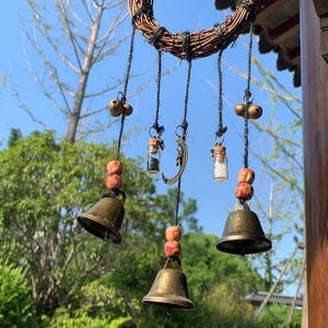 Könnte beinhalten: Ein rustikaler Windspiel mit einem geflochtenen Kranz und hängenden Elementen. Das Windspiel umfasst kleine Messingglocken, Glasfläschchen und dekorative Perlen. Der Hintergrund zeigt einen klaren blauen Himmel und üppiges grünes Laub.