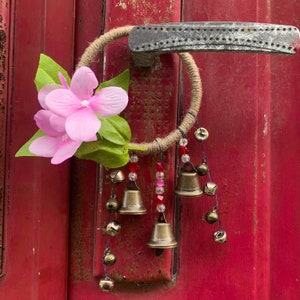 Könnte beinhalten: Eine rosa Blume mit grünen Blättern hängt an einem braunen Seilkreis mit kleinen Glocken und roten und durchsichtigen Perlen. Der Kreis ist an einem Türknauf befestigt.