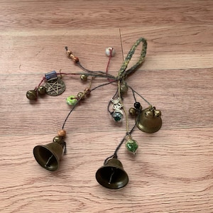 May include: A handmade charm bracelet with multiple bells, beads, and charms, including a pentagram, a crescent moon, and a small glass vial filled with green moss. The bracelet is made with a variety of materials, including leather, string, and metal.