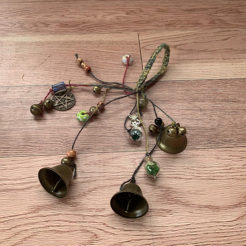 May include: A handmade charm bracelet with multiple bells, beads, and charms, including a pentagram, a crescent moon, and a small glass vial filled with green moss. The bracelet is made with a variety of materials, including leather, string, and metal.