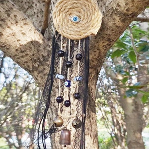 Puede incluir: Un atrapasueños tejido marrón y beige con un amuleto de ojo azul malvado colgando de una rama de árbol. El atrapasueños tiene encaje negro y cuentas, y pequeñas campanas doradas que cuelgan de la parte inferior.