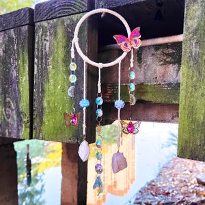May include: A white dream catcher with a pink butterfly charm, blue and clear crystal beads, and white crystal stones hanging from it.