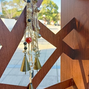 Könnte beinhalten: Ein beige-brauner Makramee-Wandbehang mit mehreren kleinen Glocken, Perlen und einem Stern-Anhänger. Der Behang ist an einer Holzlattenwand befestigt.