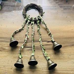 May include: A wreath made of twigs with five strands of twine with green leaves attached. Each strand has a brass bell at the end.