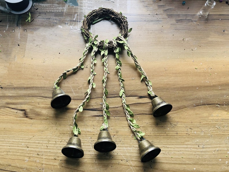 May include: A wreath made of twigs with five strands of twine with green leaves attached. Each strand has a brass bell at the end.