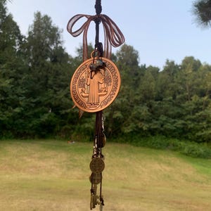 Puede incluir: Un colgante religioso de color bronce con una imagen detallada de un santo, colgado de una rama de árbol. El colgante está adornado con una cinta marrón y varios amuletos pequeños en forma de moneda. El fondo presenta un campo verde y árboles.