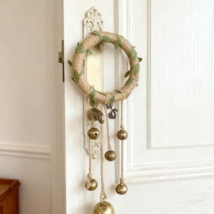 Puede incluir: Un adorno colgante para puerta con una corona envuelta en yute adornada con hojas verdes de tela. Debajo, varias campanas de latón de diferentes tamaños cuelgan de hilo de yute, creando una estética rústica. La ferretería de la puerta es visible.