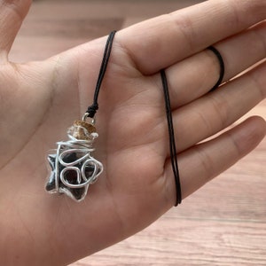 Può includere: Un ciondolo a forma di stella in vetro nero, con tappo di sughero, riempito di pietre nere e avvolto in filo d'argento, appeso a una collana con cordino nero.