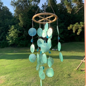 Peut inclure: Un carillon éolien suspendu avec un anneau en bambou et de multiples brins d'éléments translucides en forme de disque, de couleur sarcelle. De petites perles bleues en forme de cristal sont parsemées le long des brins. Le carillon est suspendu devant des arbres et de l'herbe verts.
