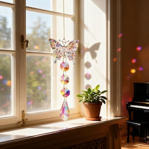 Könnte beinhalten: Ein farbenfroher Schmetterling-Sonnenfänger hängt vor einem Fenster. Der Sonnenfänger besteht aus schillernden Kristallen und hat ein Schmetterlingsdesign mit einer Kette. Unter dem Schmetterling befinden sich drei runde Kristalle und ein tropfenförmiger Kristall. Das Fenster ist weiß.