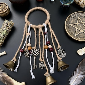 Peut inclure: Un attrape-rêves artisanal avec un anneau en jute, des brins blancs et en jute, des perles en bois et des breloques en métal. Les breloques comprennent un pentagramme, une clé et un arbre de vie. Deux cloches en laiton sont suspendues en bas.