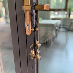 May include: A decorative door hanging featuring a black woven cord with small brass bells and a tree of life charm. The bells are of varying sizes and hang from the cord, creating a rustic and charming aesthetic.