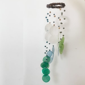 Puede incluir: Un carillón blanco, azul y verde con un anillo de madera en la parte superior y múltiples piezas redondas con forma de concha que cuelgan.