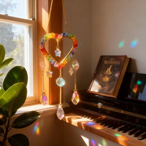 May include: A heart-shaped suncatcher with a gold frame and rainbow-colored beads. Clear crystal pendants and star-shaped charms dangle from the frame. The suncatcher hangs in front of a window, reflecting light. A book with the title "Moonbeams" is on a piano.
