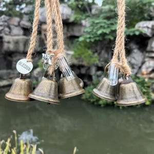 May include: A hanging wind chime featuring small, antique brass bells and clear glass vials suspended from natural rope. A small, white circular tag with the word "BLESSED" is attached. The background includes a blurred view of water and greenery.