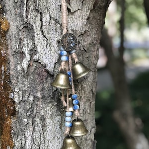 Puede incluir: Un carillón de viento con cuatro campanas de latón y cuentas azules y blancas que cuelgan de una rama de árbol. La parte superior del carillón tiene un amuleto en forma de estrella.