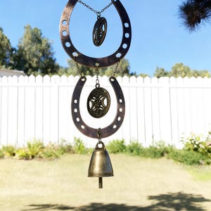May include: A bronze-colored wind chime with a horseshoe design. The chime features two horseshoe shapes, decorative oval pendants, and a bell at the bottom. The wind chime is hanging from a tree branch against a backdrop of a white fence and green grass.