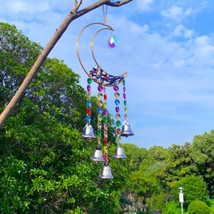 Puede incluir: Un carillón de viento en forma de media luna dorada con cuentas de cristal de colores y campanas plateadas. El carillón está colgado de una rama.