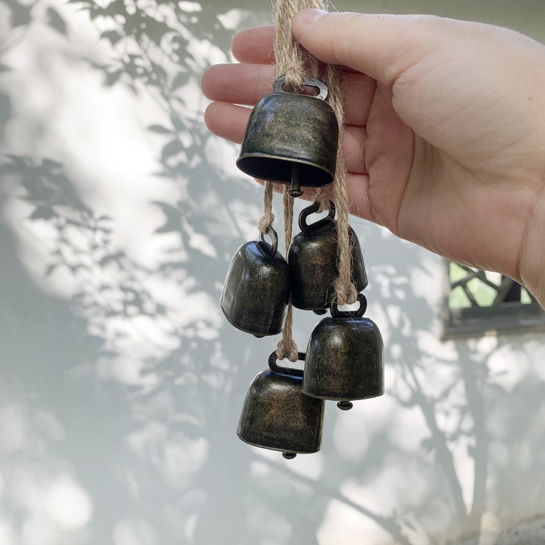 May include: A set of six small, antique-looking, silver bells hanging from a twine string. The bells are arranged in a cascading pattern, with the largest bell at the top and the smallest bell at the bottom.