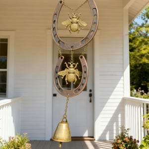 May include: A decorative wind chime featuring two horseshoe-shaped elements with a bronze finish, each adorned with a golden bee pendant. A brass bell hangs below. The wind chime is suspended by a chain, perfect for outdoor decor.