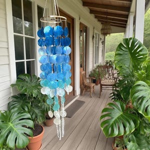 May include: A hanging wind chime with a circular top and bottom, featuring graduated blue and white shell discs. The chime is suspended by a silver chain and hangs on a porch with plants and wooden furniture.