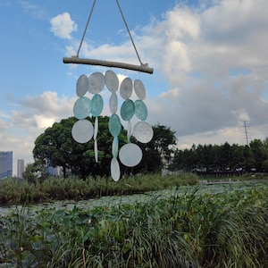 May include: A white and blue seashell wind chime hanging from a wooden dowel. The wind chime is made of multiple round seashells, some with a light blue tint and others with a white finish. The wind chime is hanging against a blue sky with white clouds.