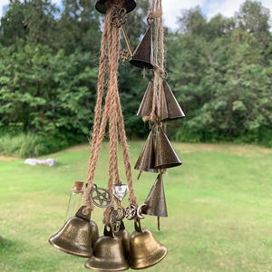 May include: A hanging wind chime featuring bronze-colored bells and charms suspended from natural rope. The chime includes various bell shapes, a small glass bottle, and decorative charms with star and key designs. The background shows green foliage.