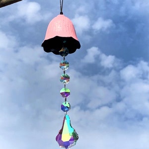 Może przedstawiać: Różowa ceramiczna dzwonnica wiatrowa z czarną obwódką i wiszącym kryształowym pryzmatem. Dzwonnica wiatrowa jest zawieszona na gałęzi na tle niebieskiego nieba z białymi chmurami.