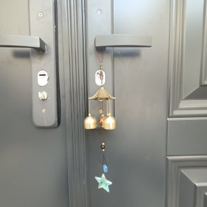 Peut inclure: Un carillon doré avec un sommet en forme de pagode et trois cloches qui pendent. Le carillon est fixé à une porte avec une poignée noire. Un charme bleu en forme d'étoile pend au bas du carillon.