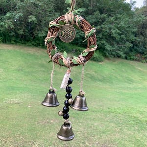 May include: A handcrafted, circular wall hanging featuring a woven grapevine wreath adorned with faux greenery and a bronze-colored pendant with a triple moon design. Below, three bronze bells and black beads hang from twine.