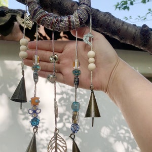 Puede incluir: Un carillón artesanal con múltiples hilos de cuentas y campanas. Los hilos presentan cuentas de madera, cuentas de vidrio y campanas de metal. La parte superior del carillón es un círculo tejido con un diseño de hoja.