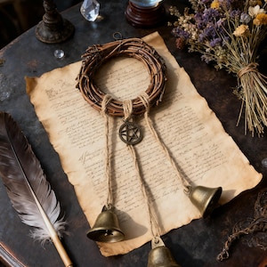 Puede incluir: Una corona rústica hecha de ramitas tejidas, adornada con un amuleto de pentagrama de bronce y tres pequeñas campanas suspendidas por hilo. La corona descansa sobre papel de pergamino envejecido, junto a una pluma y flores secas.