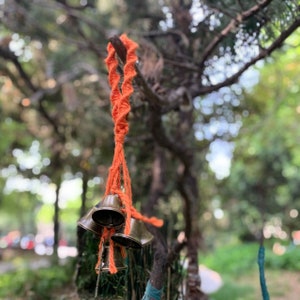 May include: A set of four small, brass bells hanging from a bright orange macrame cord. The bells are tied together with a knot and are hanging from a tree branch.