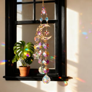 May include: A celestial-themed suncatcher with a gold-colored crescent moon and star design, adorned with colorful crystal beads and a large teardrop-shaped crystal at the bottom. The suncatcher hangs in front of a window, reflecting light.