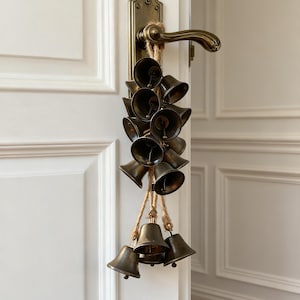 Puede incluir: Un grupo de pequeñas campanas de latón antiguo cuelgan de un tirador de puerta. Las campanas están suspendidas con cuerda natural, creando un acento rústico y decorativo. El tirador de la puerta y la ferretería circundante también son de latón.